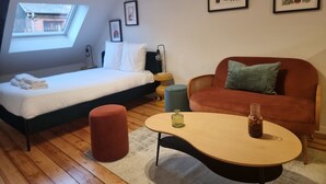 Een moderne slaapkamer met een bed, een rode poef, een ronde tafel met een glas erop, en twee ingelijste botanische prints aan de muur.