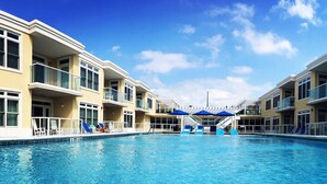Una piscina con un cielo azul despejado, un edificio de varios pisos con balcones y una zona cubierta con sombrillas.