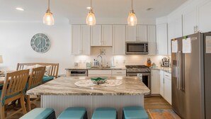 Una cocina moderna con armarios blancos, una isla central con taburetes, iluminación colgante y electrodomésticos de acero inoxidable.
