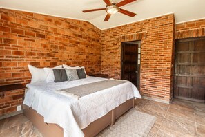 Une chambre avec un mur en briques, une porte en bois, un lit avec une literie blanche et un ventilateur de plafond.