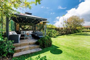 Une terrasse extérieure dotée d’un espace salon couvert, d’un escalier en pierre et d’une pelouse bien entretenue.