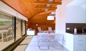 Un interior moderno con techo de madera, una zona de comedor con mesa y sillas, una cocina con fregadero y vistas amplias a través de grandes ventanas.