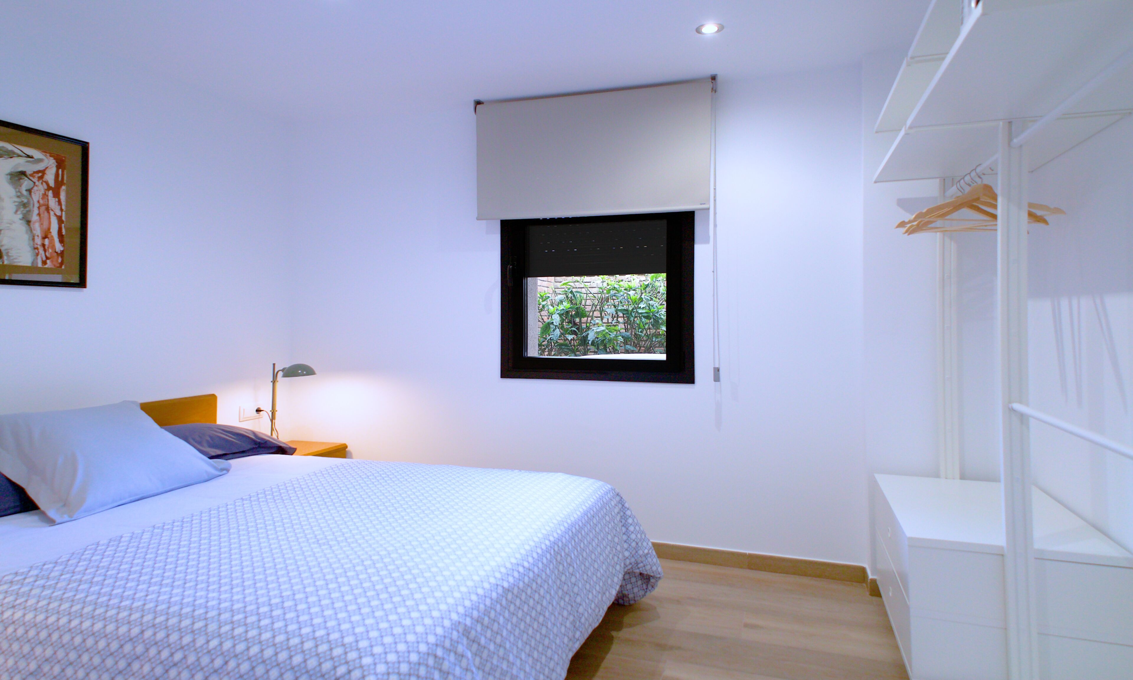 A bedroom with a bed, a nightstand, a lamp, a window with a view of greenery, and a wardrobe.