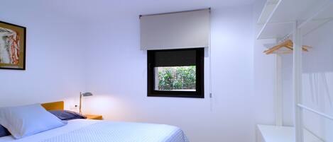 A bedroom with a bed, a nightstand, a lamp, a window with a view of greenery, and a wardrobe.