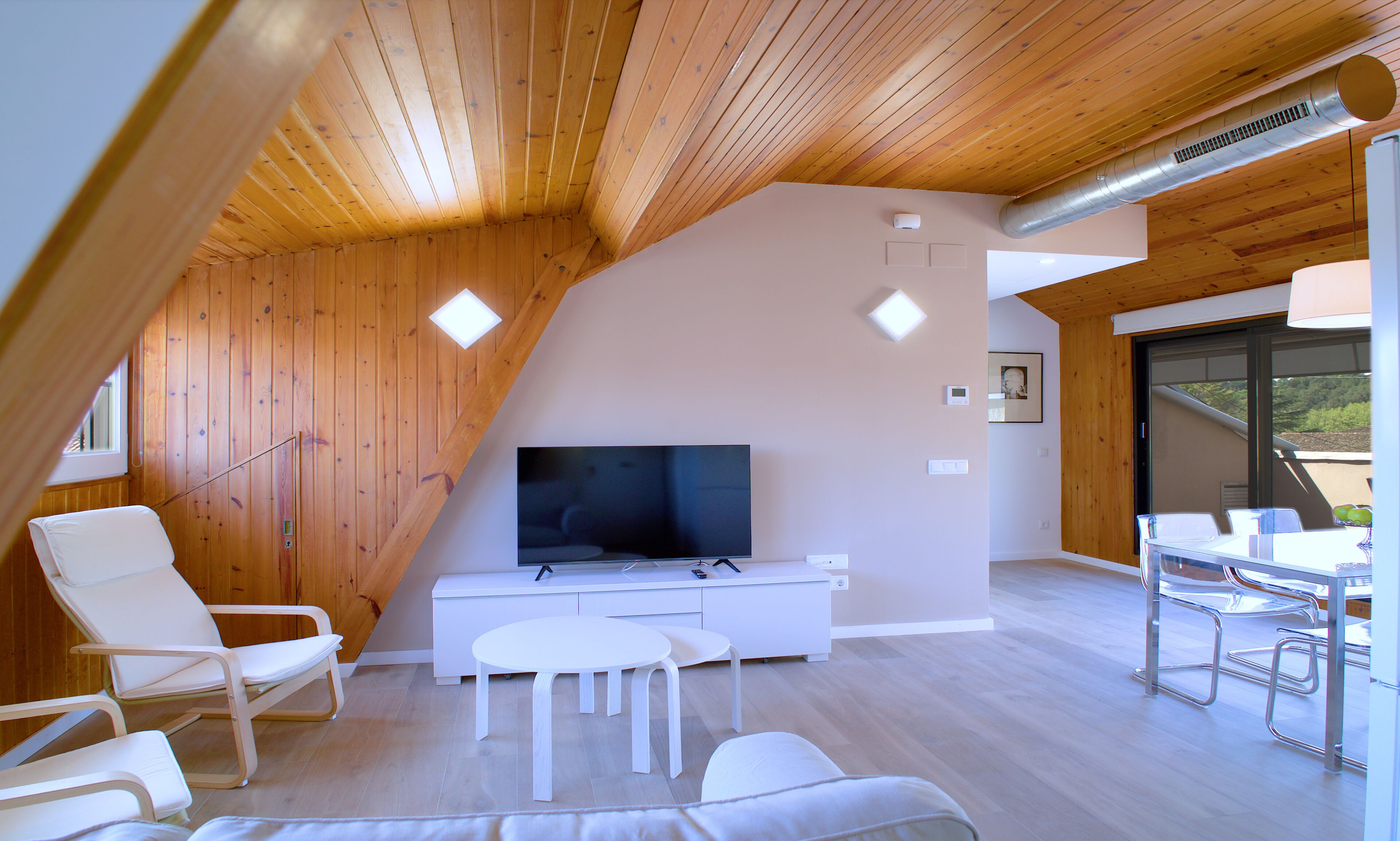 A modern interior with a sloped ceiling, wooden walls, a flat-screen TV, white furniture, and a dining area.