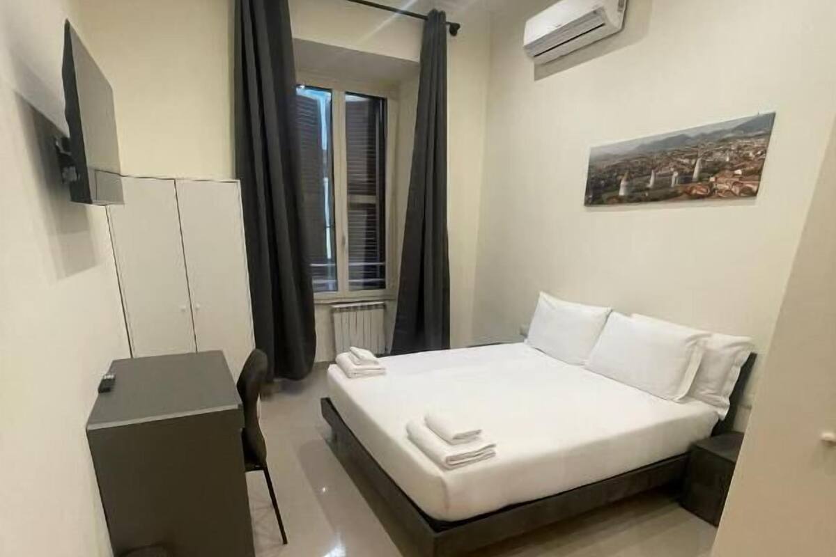Una habitación de hotel con cama, escritorio, silla, cesto de basura y ventana con cortinas.