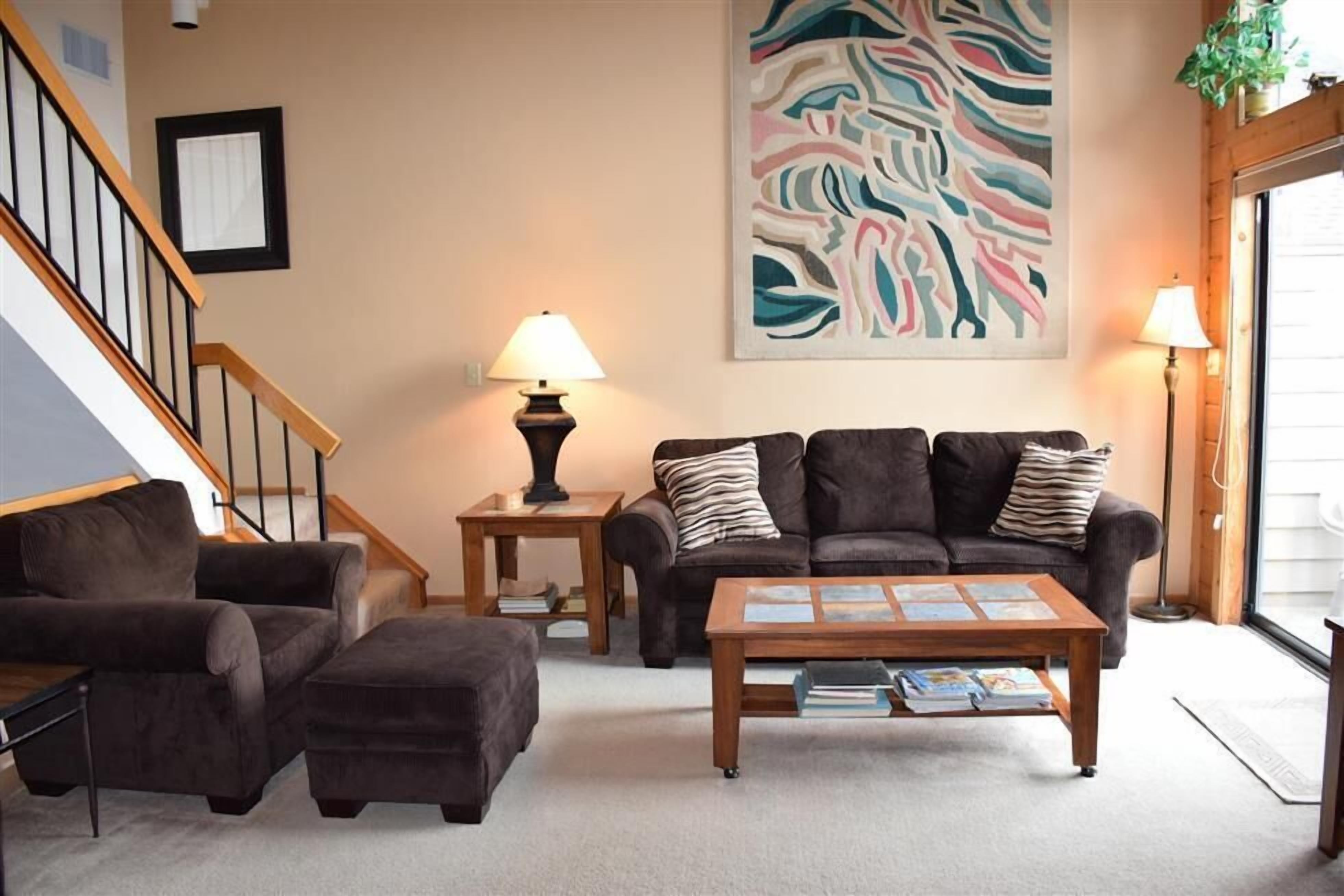 A living room with a sofa, ottoman, coffee table, and two side tables with lamps. A staircase is visible in the background.