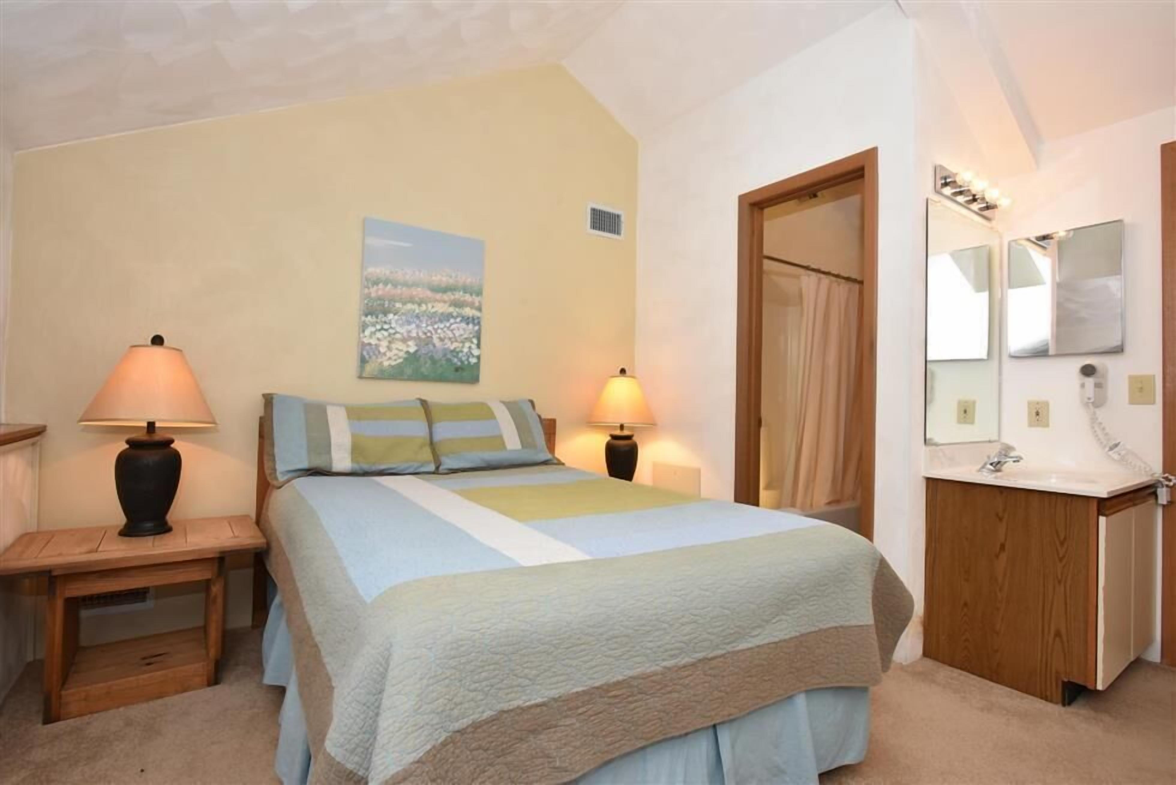 Ein Schlafzimmer mit einem Bett, zwei Nachttischlampen, einem Nachttisch, einer Holztür, einem Badezimmer mit Waschbecken und Spiegel sowie einem Bild an der Wand.
