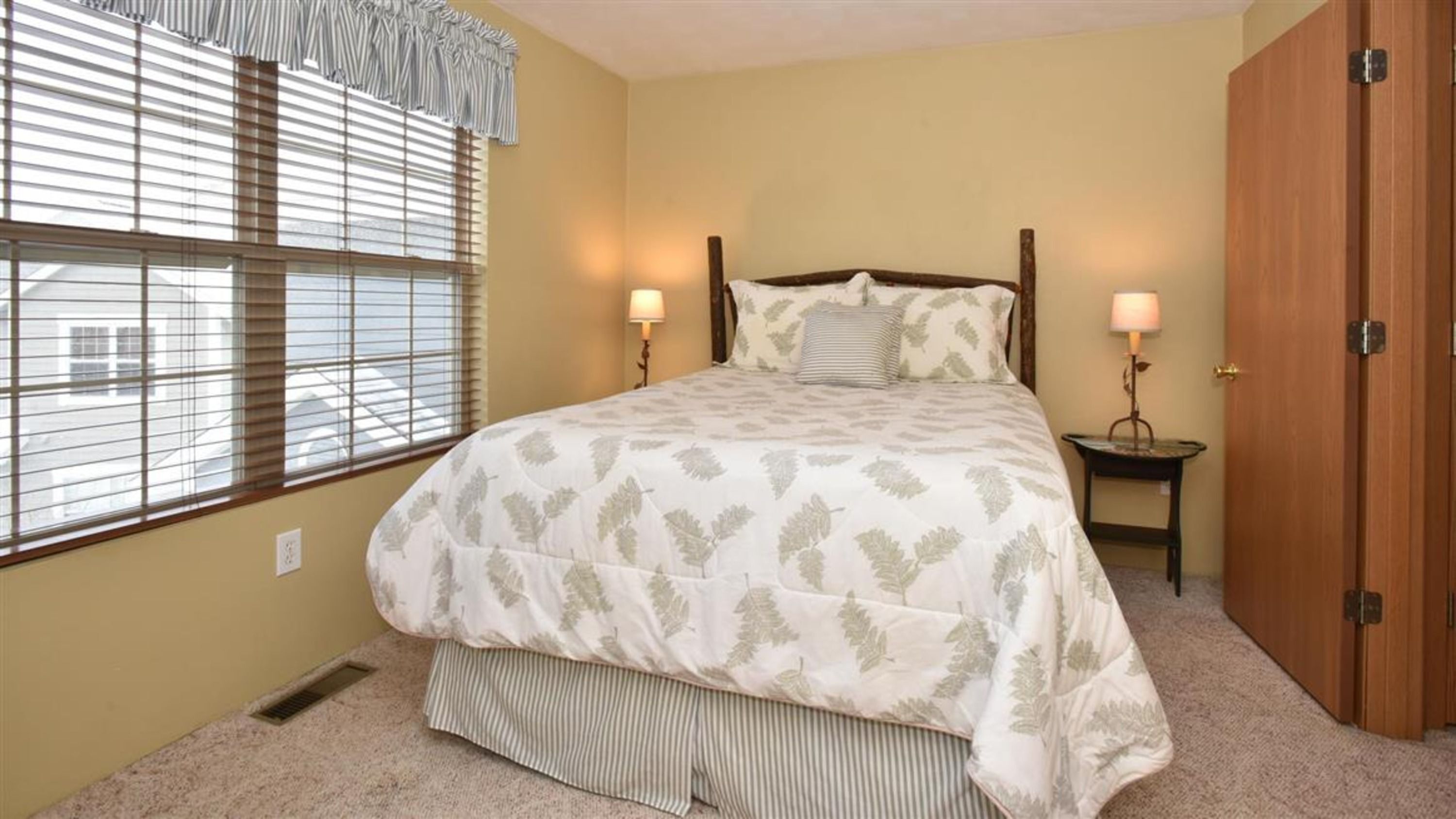A bedroom with a bed, two bedside lamps, a window with blinds, a wooden wardrobe, and a carpeted floor.