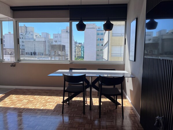Un coin repas avec une table et des chaises, de grandes fenêtres donnant sur la ville et un radiateur fixé au mur.