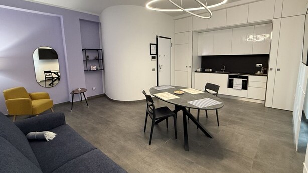 Una sala de estar moderna con sofá, una mesa de comedor con sillas, una zona de cocina y un espejo circular en la pared.