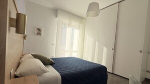 Un dormitorio moderno con cama, mesita de noche, lámpara de pared y ventana con cortinas translúcidas.