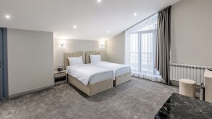 Une chambre d’hôtel moderne dotée d’un grand lit, de tables de chevet, d’un bureau et offrant une vue sur la ville grâce à de grandes fenêtres.