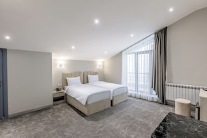 Habitación de hotel moderna con una cama grande, mesitas de noche, un escritorio y vistas de la ciudad a través de amplias ventanas.