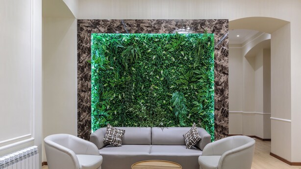 Un salon moderne avec un canapé gris, deux fauteuils et une table basse en bois. Un grand mur vert, agrémenté d’un motif de feuilles et éclairé, se trouve derrière le canapé.