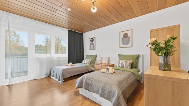 A bedroom with two beds, a wooden ceiling, a large window with curtains, and a vase with flowers on a nightstand. - Tina in Buchen (Buchen)