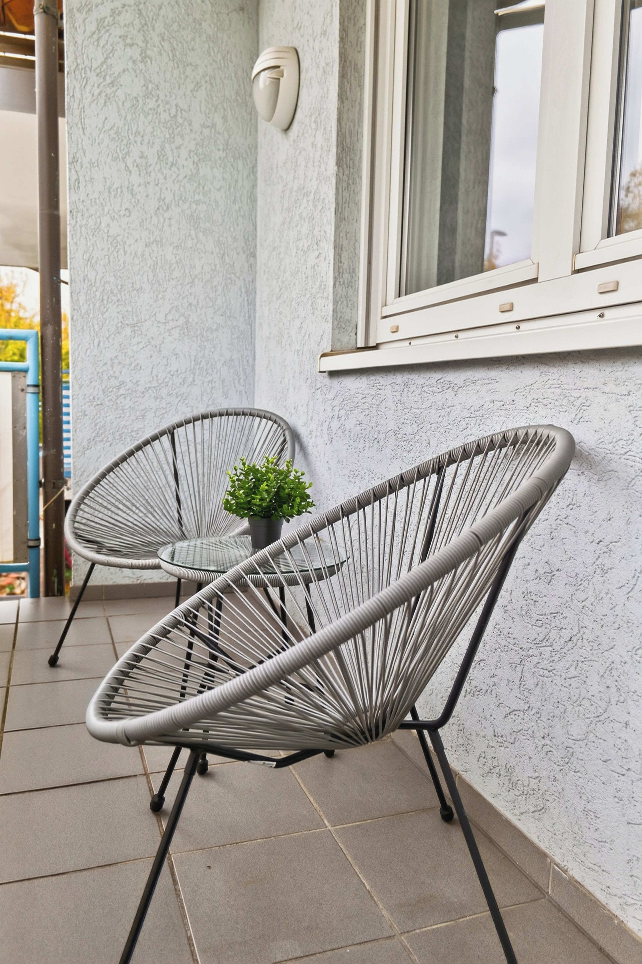 Deux fauteuils en osier gris, avec une petite plante en pot, sont placés sur une terrasse carrelée, juste à côté d’un mur blanc à la texture particulière et d’une fenêtre équipée de stores.