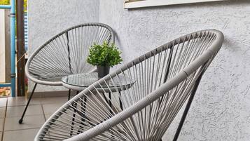 Deux fauteuils en osier gris, avec une petite plante en pot, sont placés sur une terrasse carrelée, juste à côté d’un mur blanc à la texture particulière et d’une fenêtre équipée de stores.