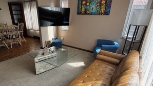 Een woonkamer met een bruine leren bank, een flatscreen televisie, een schilderij aan de muur en een eethoek met stoelen.