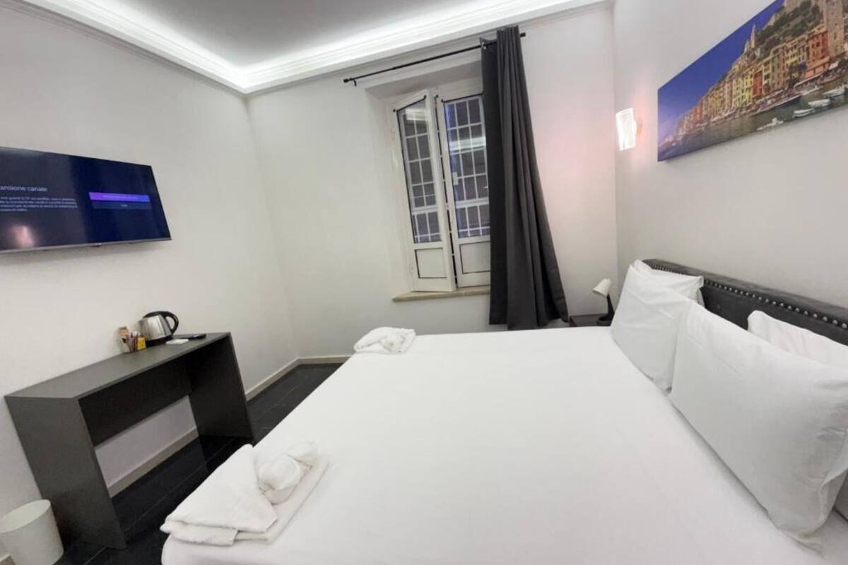 Habitación de hotel con una cama grande, un televisor de pantalla plana montado en la pared, un escritorio pequeño con una tetera y una ventana con cortinas.