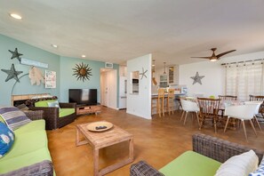 A spacious living room with a television, green sofas, and a dining area with wooden chairs and a table. - Mission Bay Waterfront | Patio Steps to the Sand (San Diego)