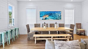 Un comedor con mesa de madera, sillas de mimbre y un banco. Hay dos ventanas con persianas blancas, y un cuadro de una ballena en la pared.