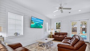 Amplia sala de estar con paredes de madera blanca, ventilador de techo, televisor de pantalla plana grande, sofás de cuero y vista a una piscina a través de puertas de vidrio.