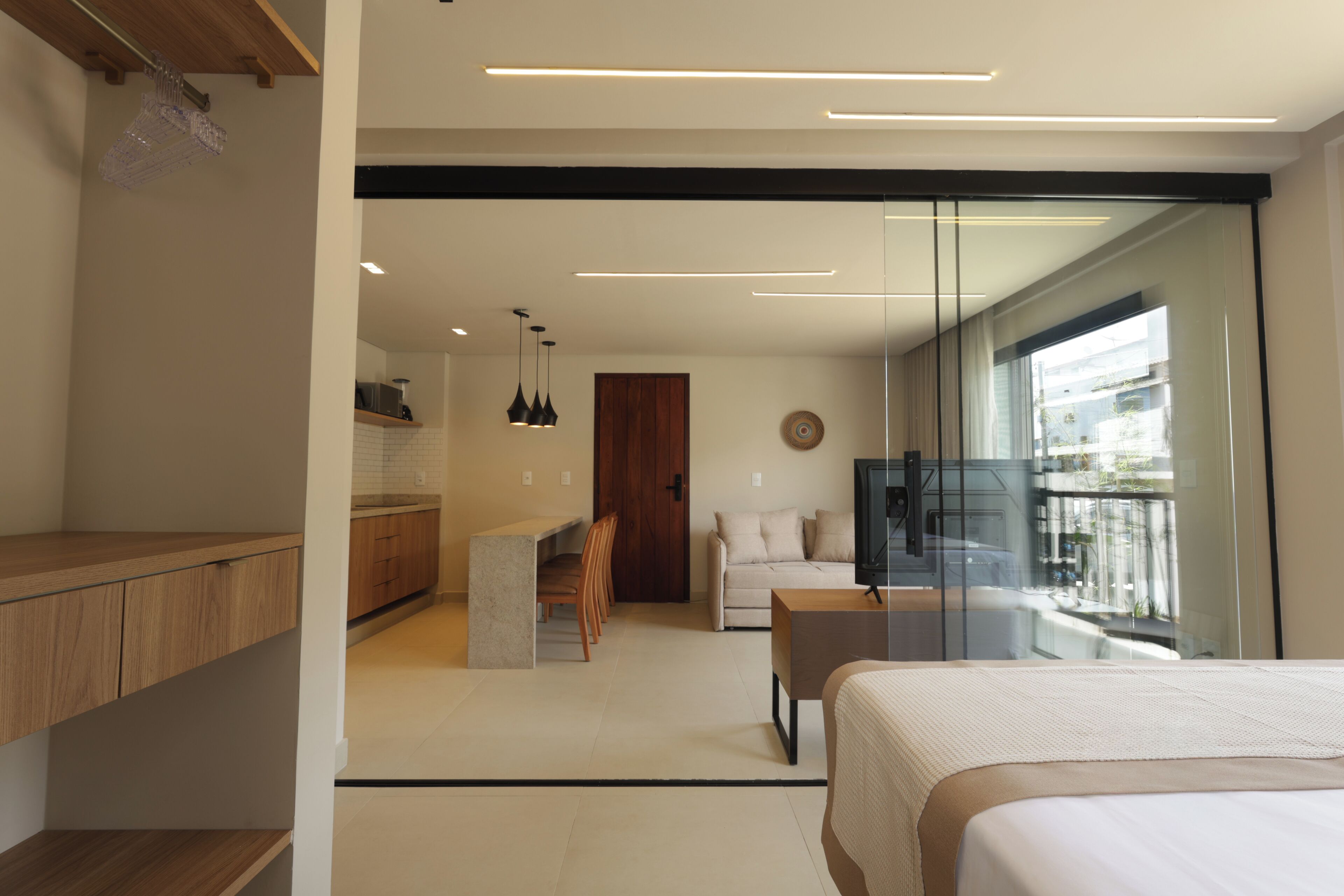 A modern hotel room with a large bed, a seating area with a sofa and chairs, a kitchen area, and a balcony with a view.