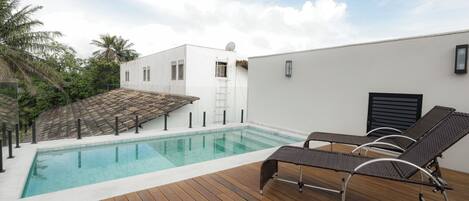 A rooftop pool with wooden deck and lounge chairs, surrounded by a white building and palm trees.