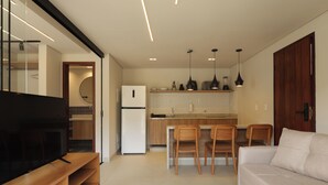 A modern kitchen with a built-in oven, a dining area with wooden chairs, a TV on a wooden stand, and a sofa. - Jubarte Flats  (Itacaré)
