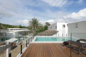 Una piscina en la azotea con terraza de madera, barandilla de vidrio y vistas a edificios y palmeras circundantes.