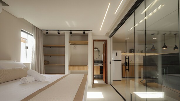 A modern hotel room with a large bed, a glass wall, and a kitchen area visible through the glass. - Jubarte Flats  (Itacaré)