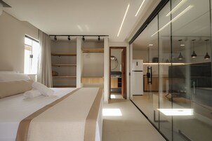 Una habitación de hotel moderna con una cama grande, una pared de vidrio y una zona de cocina visible a través del vidrio.