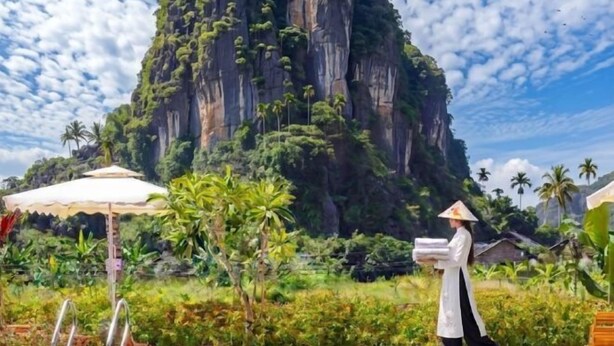 A pool area with a large rock formation, palm trees, and a person carrying towels. - Trang An Ecorest Resort & Spa (Hoa Lu District)