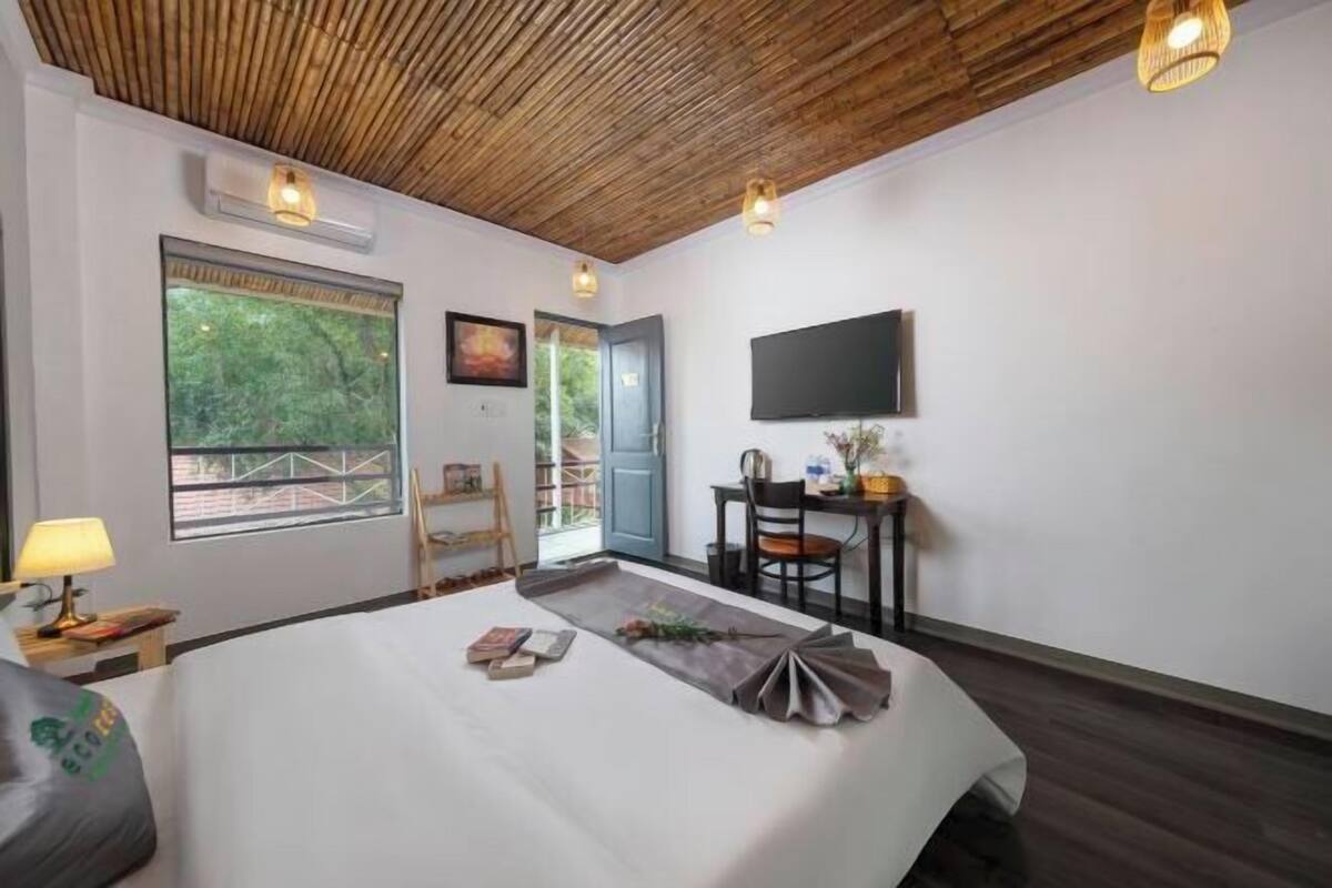 A bedroom with a bed, a desk, a chair, a TV, and a window with a view of greenery.