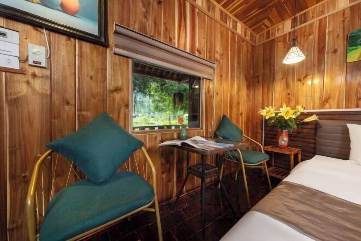 A wooden-paneled room with a bed, two chairs, a small table, and a window with a view of greenery.