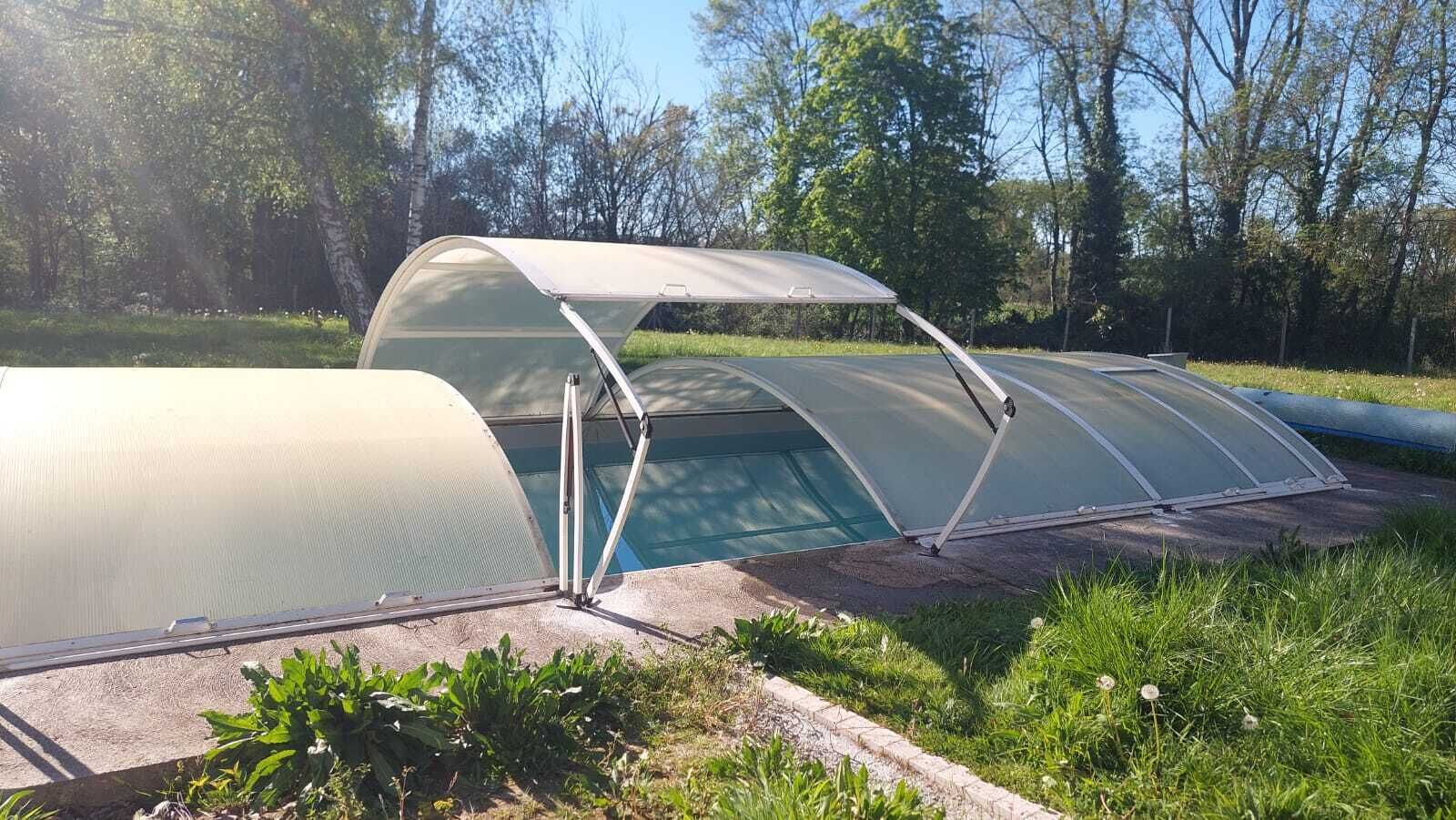 Una estructura invernadero con techo transparente y estructura metálica, ubicada en un área con césped y árboles al fondo.
