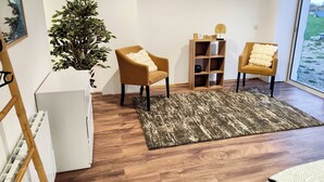 Una sala de estar moderna con dos sillones, una estantería de madera, una planta grande en maceta y vistas al exterior a través de un amplio ventanal.