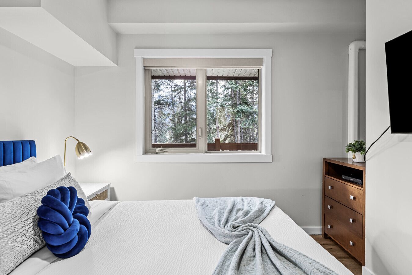 Quarto com cama, mesa de cabeceira, penteadeira, janela com vista para árvores e luminária.