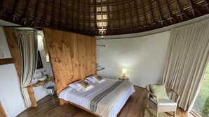Un dormitorio con cabecera de madera, cama con ropa de cama blanca, mesita de noche con lámpara, una silla de mimbre y vistas al verdor exterior.