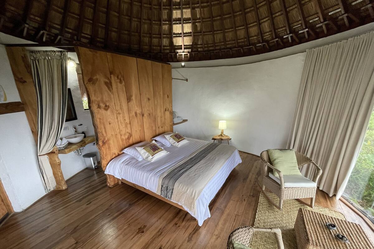 A bedroom with a wooden headboard, a bed with white bedding, a bedside table with a lamp, a wicker chair, and a view of greenery outside.