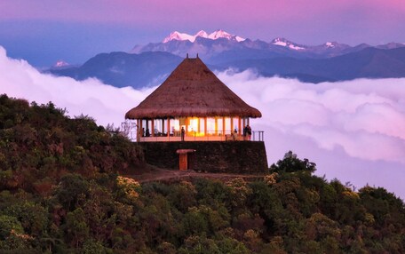 A thatched-roof hut with a wooden door, surrounded by lush greenery and overlooking a mountain range.