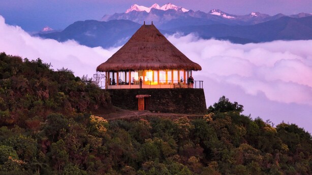 Una cabaña con techo de paja y puerta de madera, rodeada de exuberante vegetación y con vistas a una cordillera.