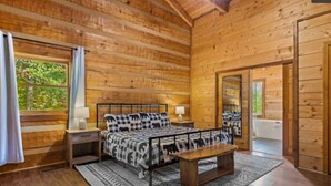 A wooden cabin bedroom with a bed, bedside tables, a bench, and a window with curtains. - The Smoky Bear: View, Hot Tub, Pool & MORE! (Gatlinburg)