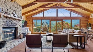 A cozy cabin interior with a stone fireplace, wooden ceiling beams, a large window with a view, and a seating area with a table and chairs. - The Smoky Bear: View, Hot Tub, Pool & MORE! (Gatlinburg)