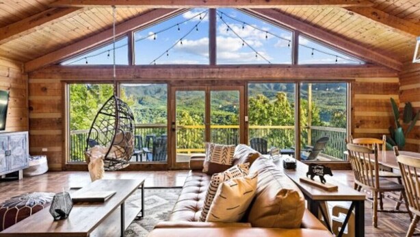 A spacious loft-style room with a high ceiling, large windows offering a view of the outdoors, a cozy seating area with a sofa and chairs, and a dining table with chairs. - The Smoky Bear: View, Hot Tub, Pool & MORE! (Gatlinburg)