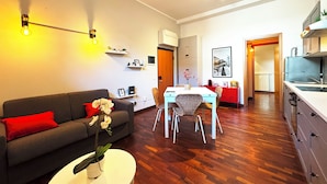 Een moderne woonkamer met een bank, een eettafel en stoelen. Er is een keukenhoek met kastjes en een gootsteen.