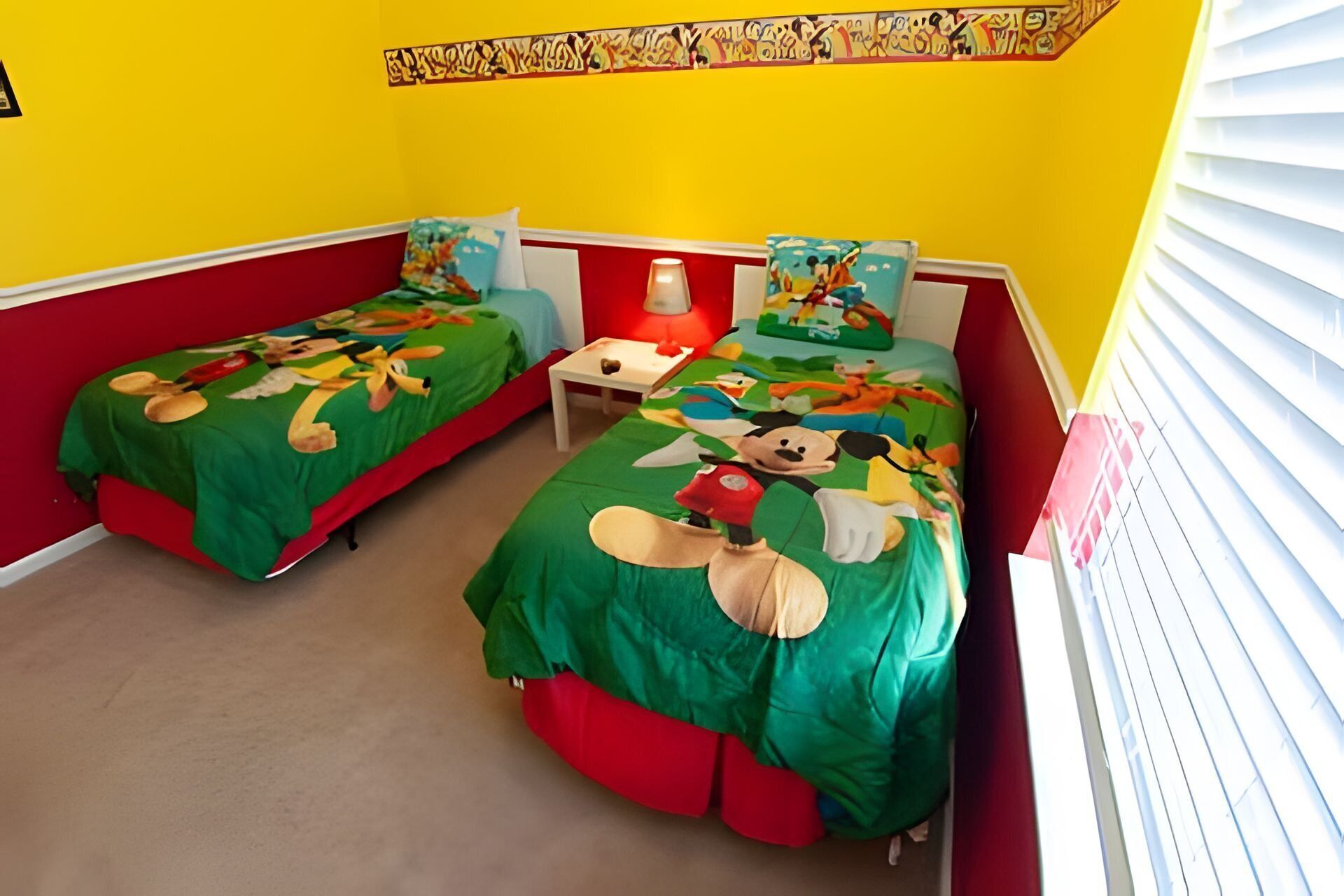 A room with two beds, each covered with green sheets featuring cartoon characters, a small table, and a lamp.