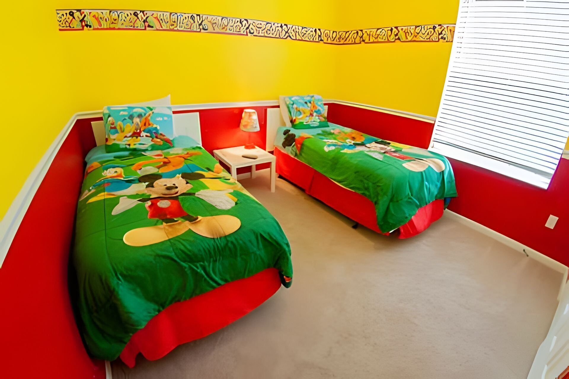 Een kamer met twee bedden, elk met een kleurrijk sprei met tekenfilmfiguren. Er is een kleine tafel en een raam met zonwering.