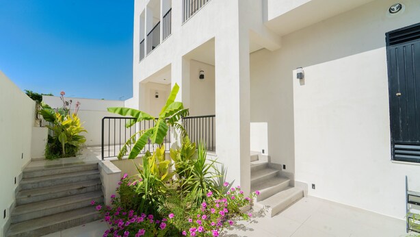 Ein modernes Gebäude mit weißer Fassade, einer Treppe, die zu einem Balkon führt, und verschiedenen Pflanzen.